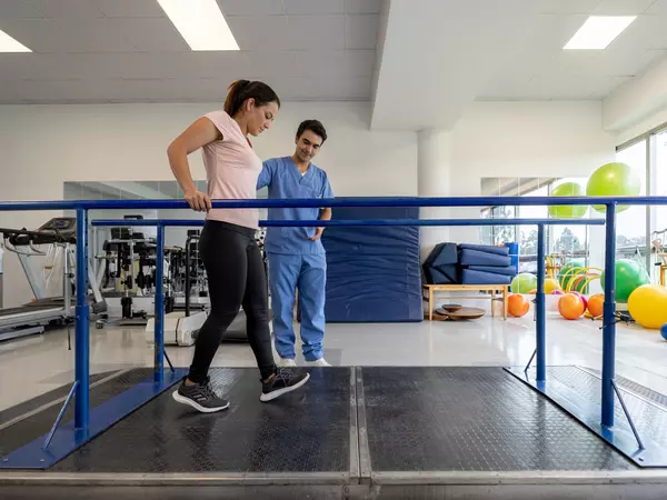 Latin American woman in physical therapy walking on the parallel bars with the assistance of his therapist.